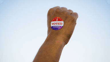 A hand with an "I Voted Today" sticker against a clear sky backdrop